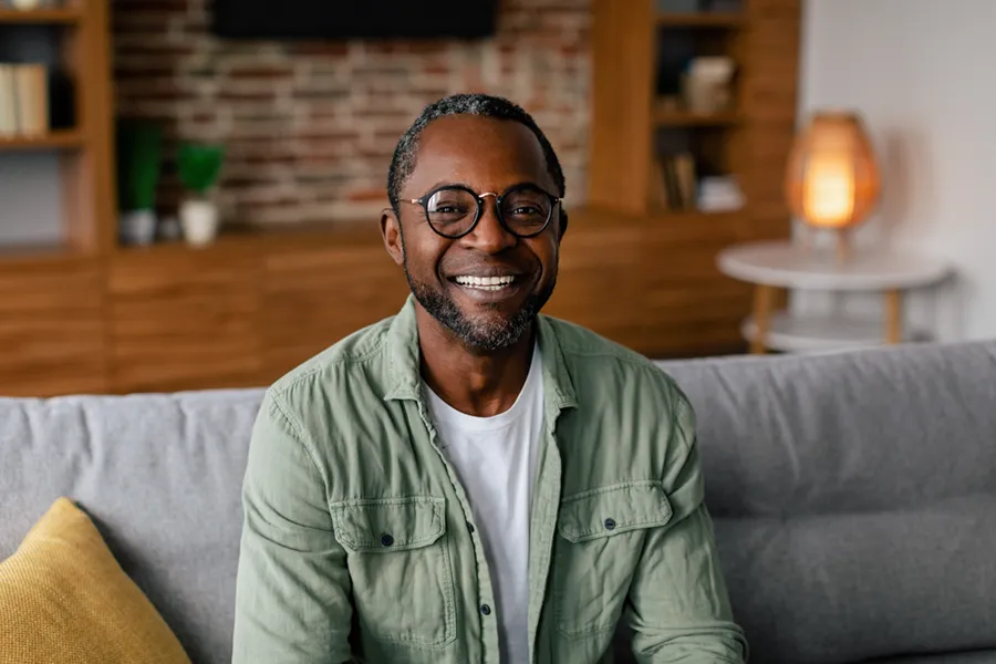 A middle aged black man in a green button up shirt sits on his couch smiling, happy with his treatment for andropause and low testosterone from The Cove Concierge Medicine in Denver.