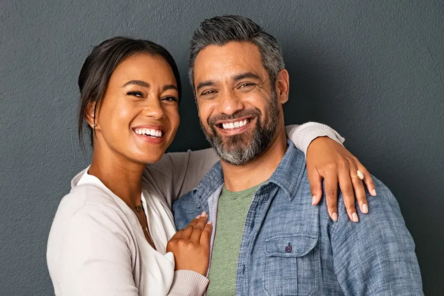 A middle-aged couple with their arms around each other standing against a gray background and smiling, happy with the bioidentical hormone therapy they received from The Cove Concierge Medicine in Denver.