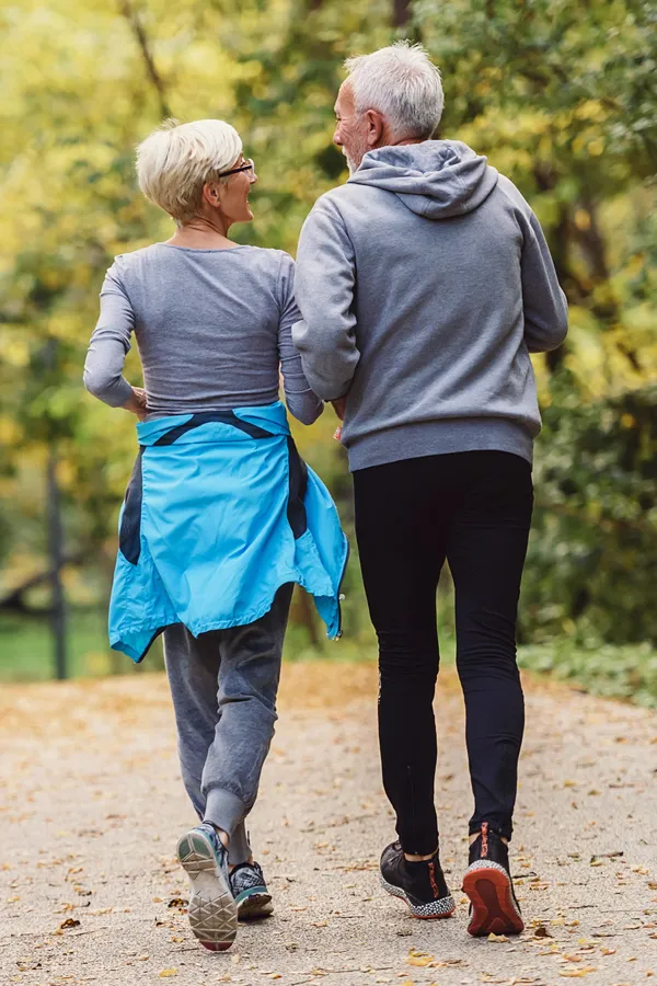 A mature couple in workout clothes walk briskly on a trail. Get a cancer screening from The Cove Concierge Medicine in Denver.