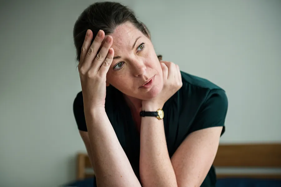 A woman in a dark blue dress sits on her bed holding her head in her hands with an exhausted expression. Get help for chronic fatigue syndrome from The Cove Concierge Medicine in [AERA].