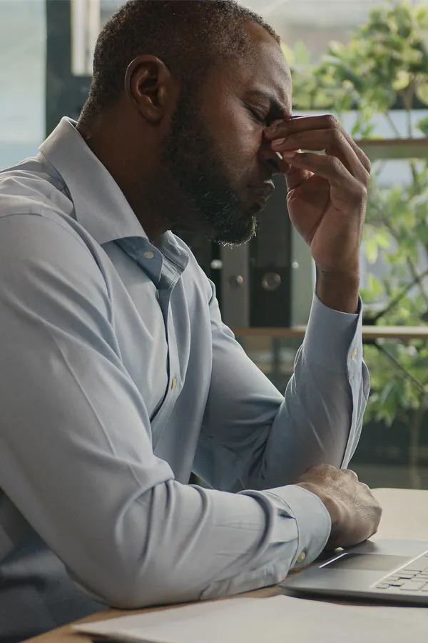 A black man rubbing the top of his nose at his desk, in need of chronic fatigue treatment from The Cove Concierge Medicine in Denver.