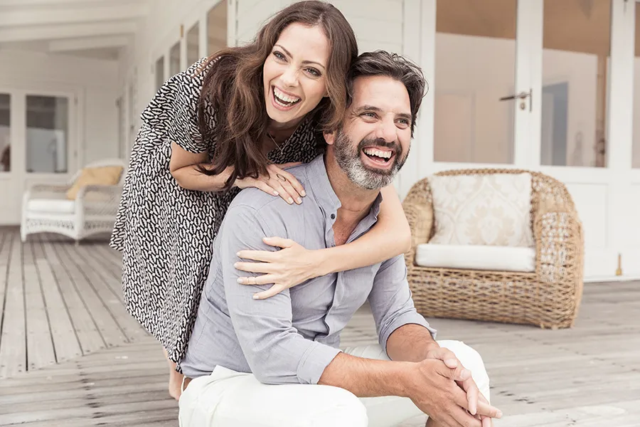 A middle-aged couple on a wooden porch with wicker furniture; the woman is playfully hugging the man from behind. They are benefiting from the concierge care services from The Cove Concierge Medicine in Denver.