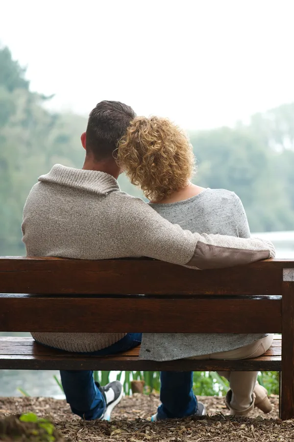 A couple in sweaters sit arm and arm on a bench looking at a calm lake. Get a proactive health screening from The Cove Concierge Medicine in Denver.