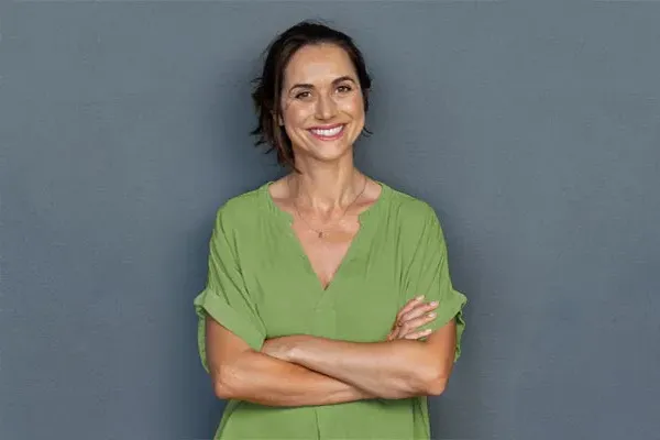 A woman in a green shirt stands smiling against a gray-blue wall, pleased with her hormone therapy from The Cove Concierge Medicine in Denver.