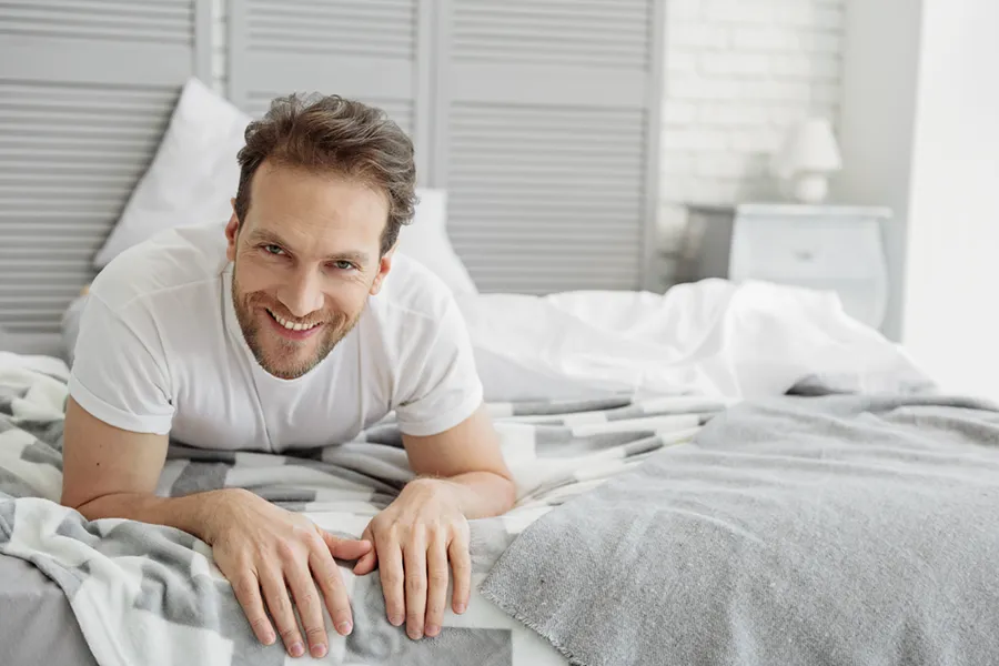 A dirty-blonde haired man lays on his stomach in bed smiling. Schedule treatment for low libido in men from The Cove Concierge Medicine in Denver.