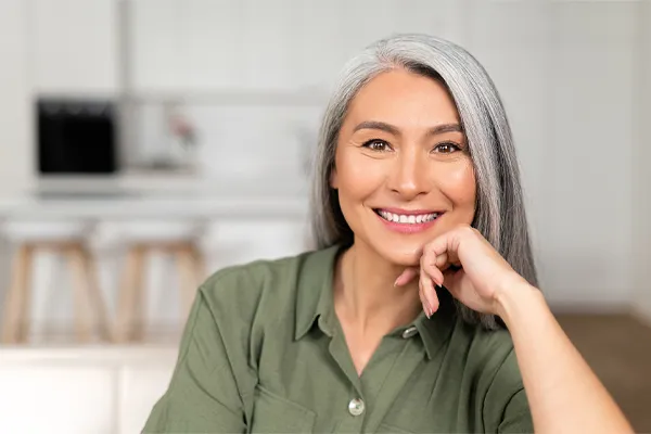 An asian woman with long gray hair and a green shirt sitting and smiling on her couch, happy with her menopause care from [NAME} in Denver.