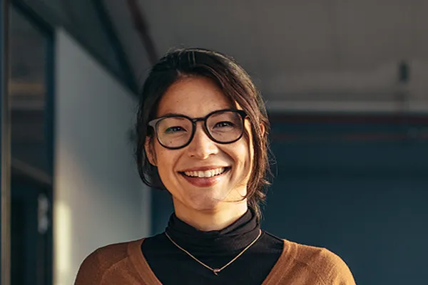 A brunette woman with glasses smiles in a gray-blue walled office. Get metabolic syndrome from The Cove Concierge Medicine in Denver.
