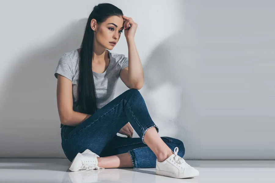 A young brunette woman in a gray t-shirt and jeans sits against a gray wall with her head resting on her hand and a distressed expression. Get help for Polycystic Ovarian Syndrome (PCOS) from The Cove Concierge Medicine in Denver.