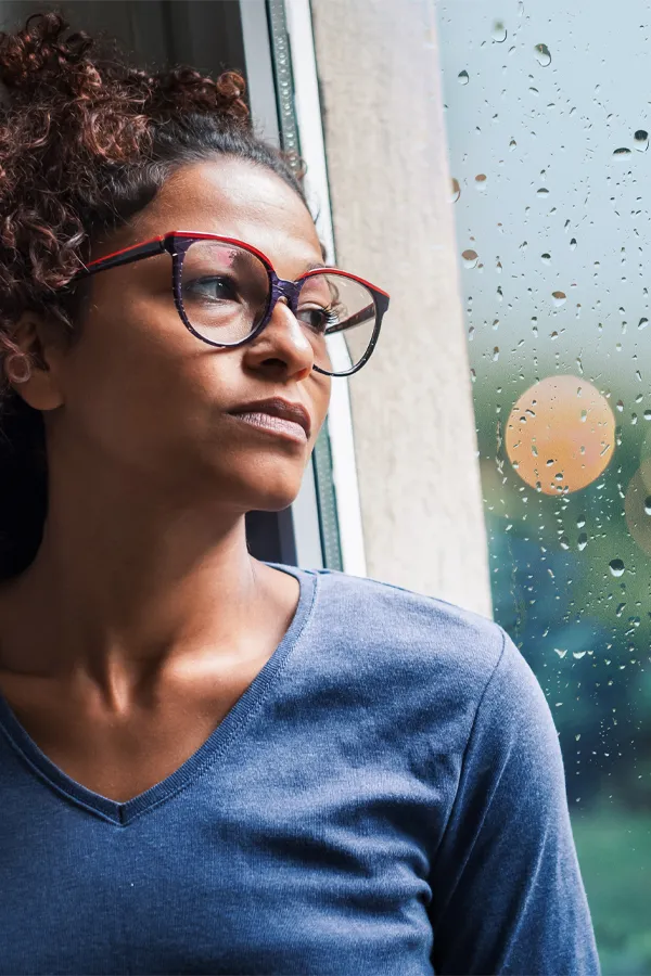 A black woman with with a somber expression and glasses and a gray shirt stares out the window on a rainy day. Get treatment for Premenstrual Dysphoric Disorder from The Cove Concierge Medicine in Denver.