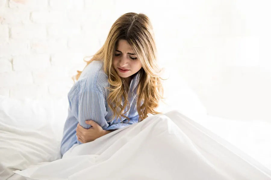A woman with long blonde hair sits in her bed holding her stomach in pain, considering getting care for PMS from The Cove Concierge Medicine in Denver.