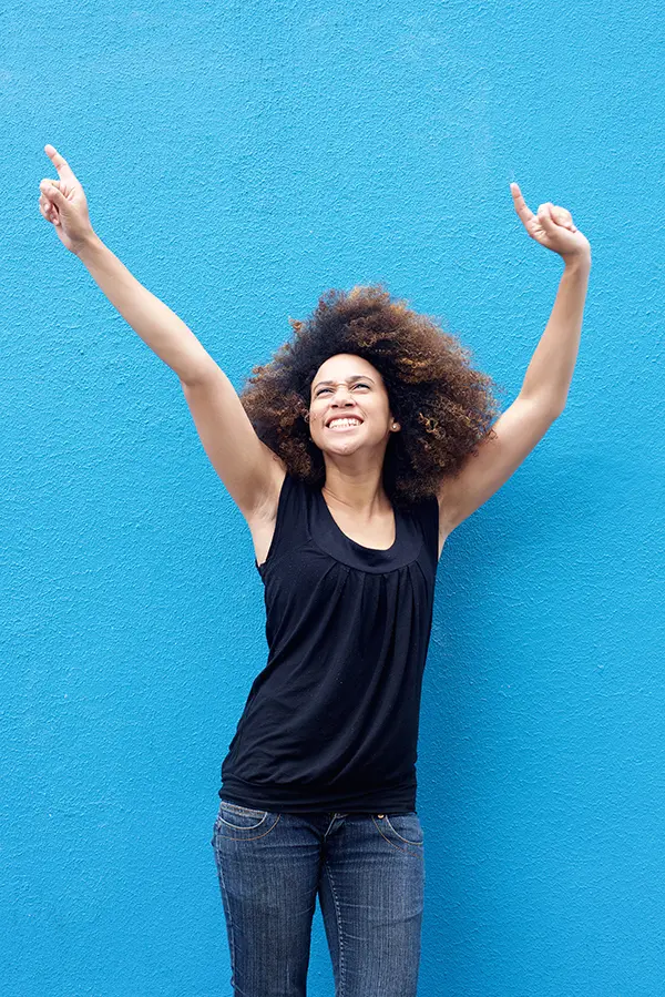 A woman in a dark blue tank top standing in front of a bright blue wall, raising her arms in celebration of relief from PMS from The Cove Concierge Medicine in Denver.