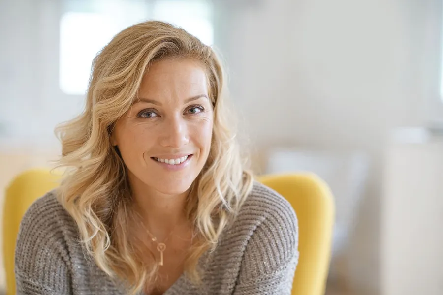 A blonde woman wearing a a gray sweater sits inside in a yellow chair, smiling and happy with her perimenopause treatment from The Cove Concierge Medicine in Denver.