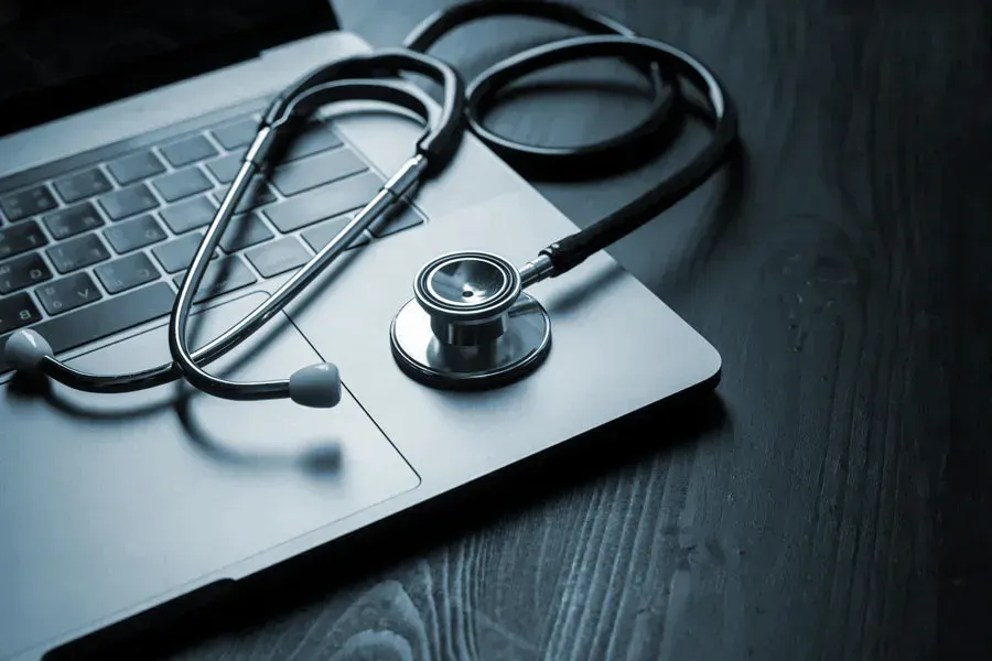 An open laptop with a stethoscope lying on it on a wooden table, symbolizing telemedicine care from The Cove Concierge Medicine in Denver.